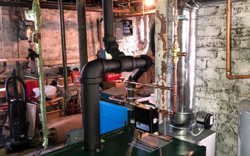 Basement boiler system with black and copper piping, a vent pipe, and surrounding storage in an industrial setting.