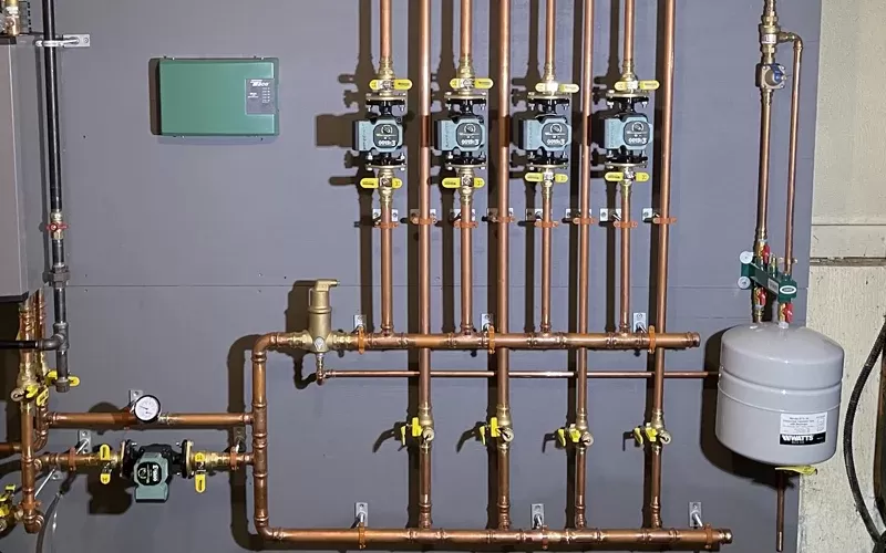 Residential heating system with copper pipes, multiple control valves, a circulation pump, and a gray expansion tank mounted on a gray wall.