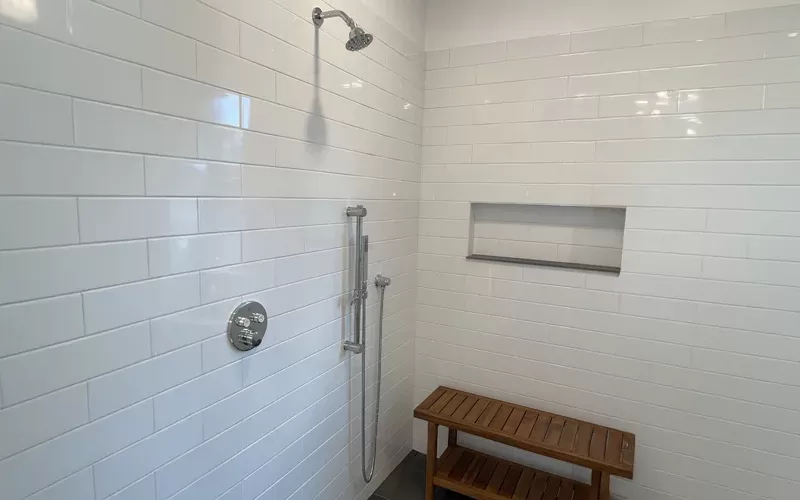 Minimalist shower with white subway tile walls, a rainfall showerhead, a handheld shower fixture, a recessed shelf, and a wooden bench.