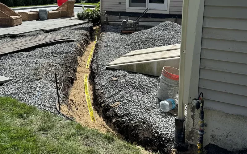 Outdoor trench near a house with a caution tape marking buried utility lines, gravel surrounding the area, and patio construction materials nearby.