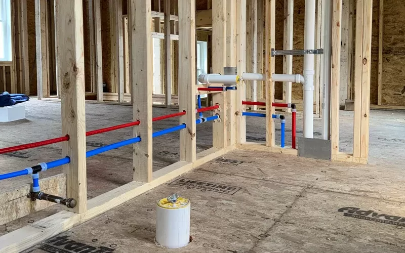 Framed walls in a construction site with red and blue water supply lines and white PVC drainage pipes installed.